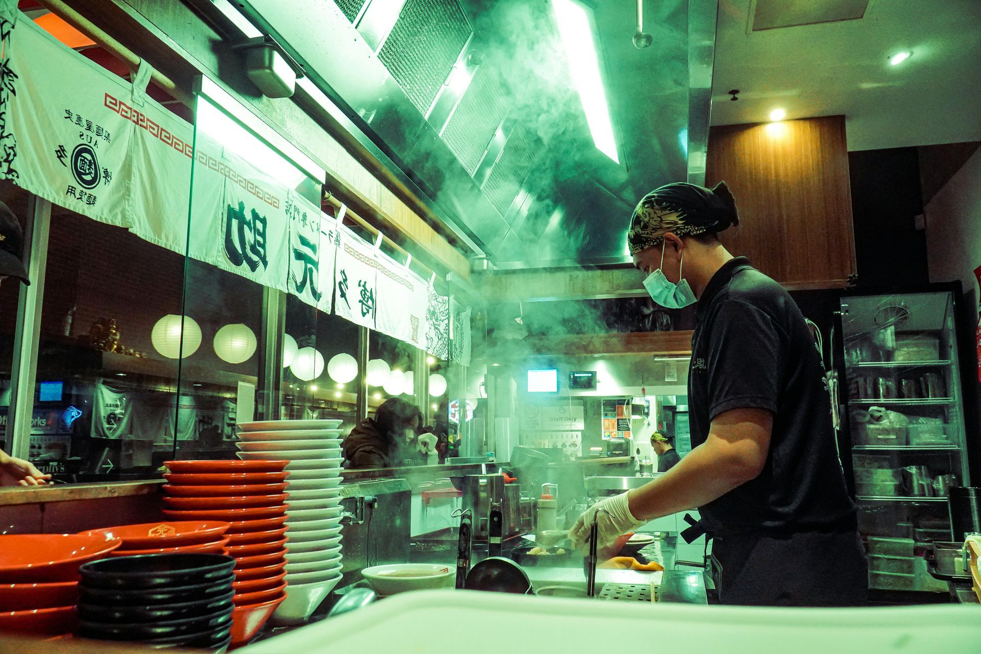 Chef in a restaurant kitchen, wearing mask and gloves, cooking near steam and stacked plates.