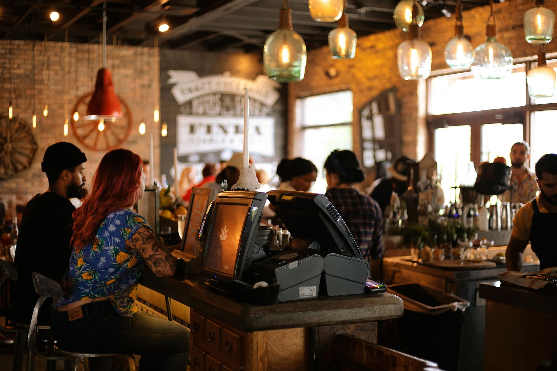 Interior of a restaurant with people seated at a bar and around tables. Overhead lights illuminate the space.