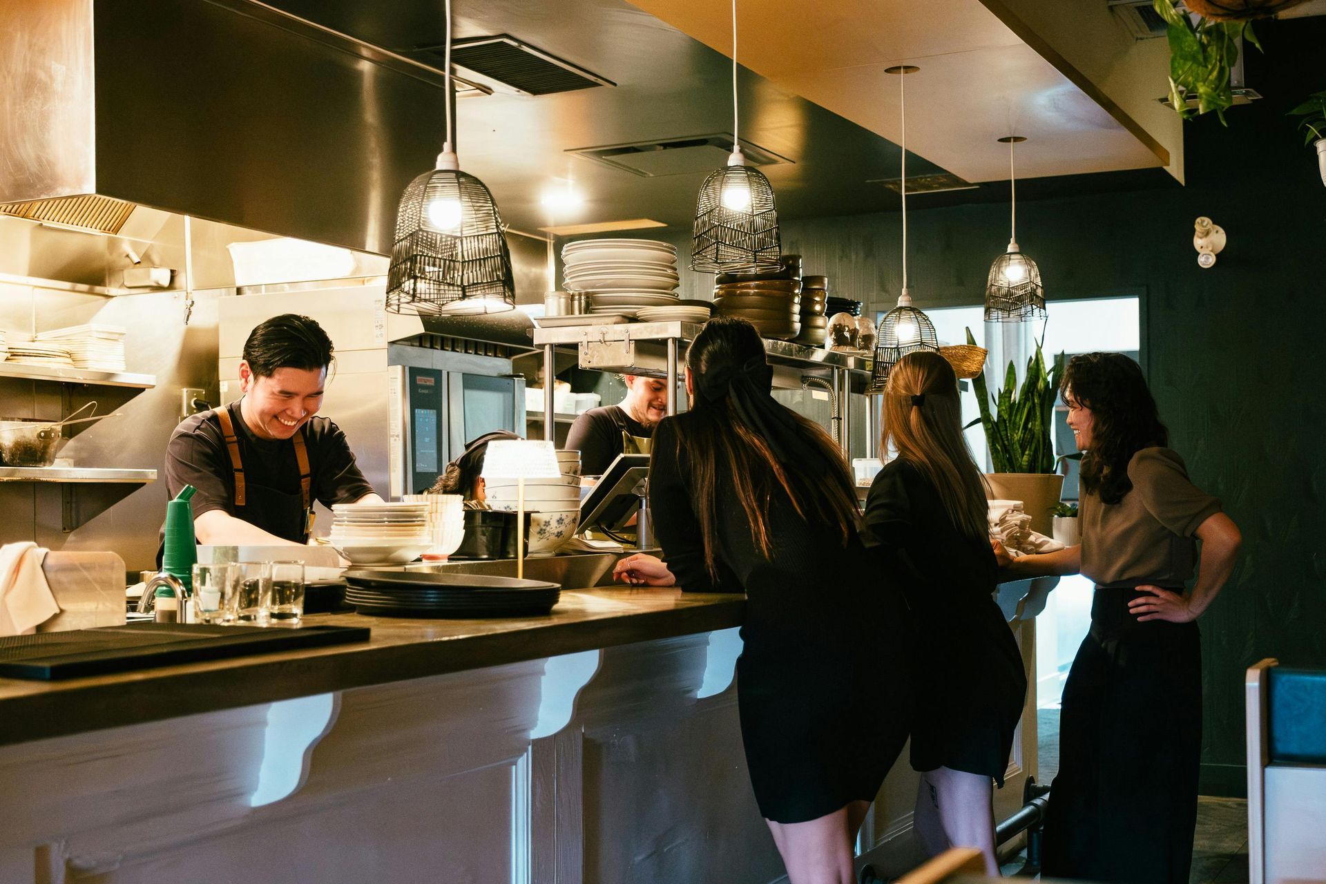 Restaurant interior: Chef at counter, laughing, serving customers, other staff in background.