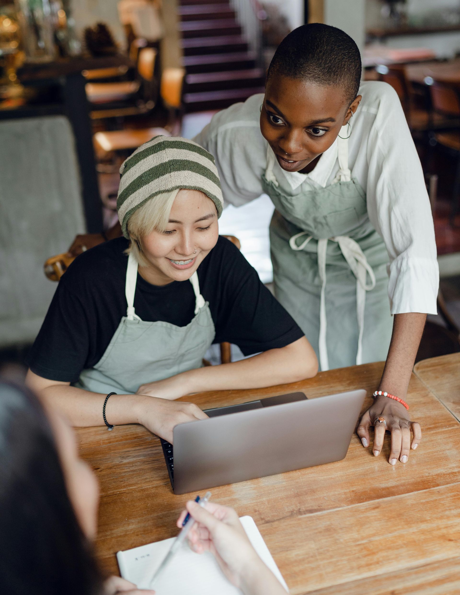 Two people in aprons look at a laptop, one leans over the other. Wooden table, cafe setting.