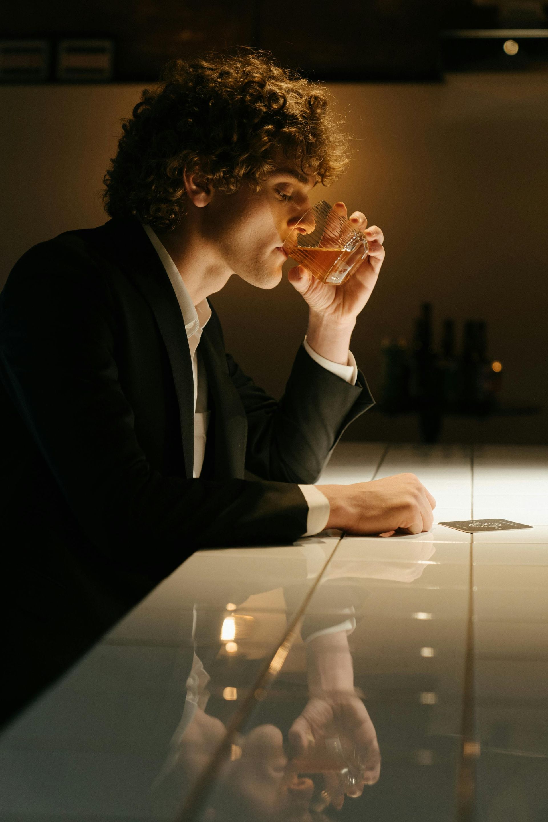Man in suit sips a drink at a bar; dim lighting, reflective white countertop.