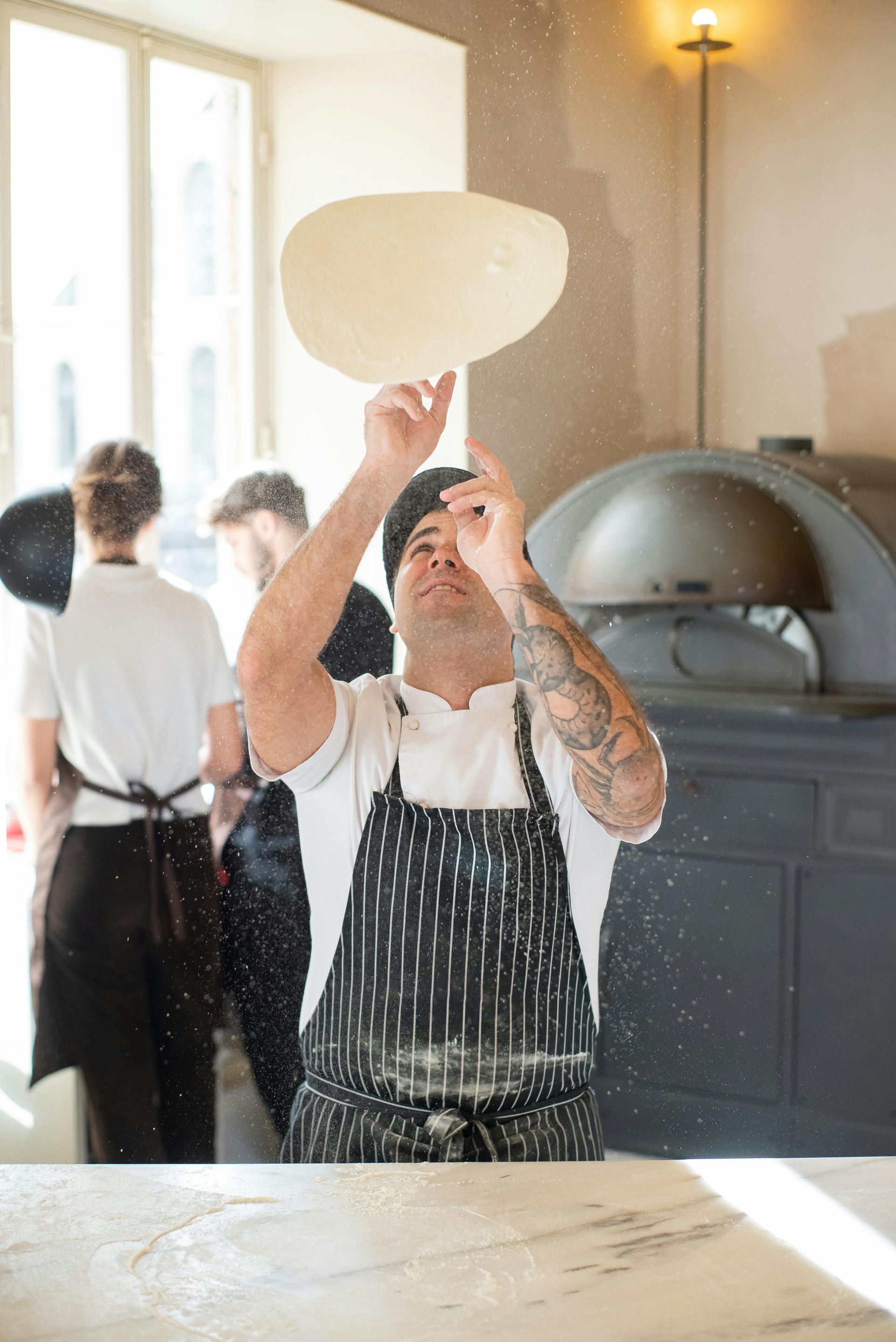 Chef tossing pizza dough, kitchen with ovens and staff in the background.