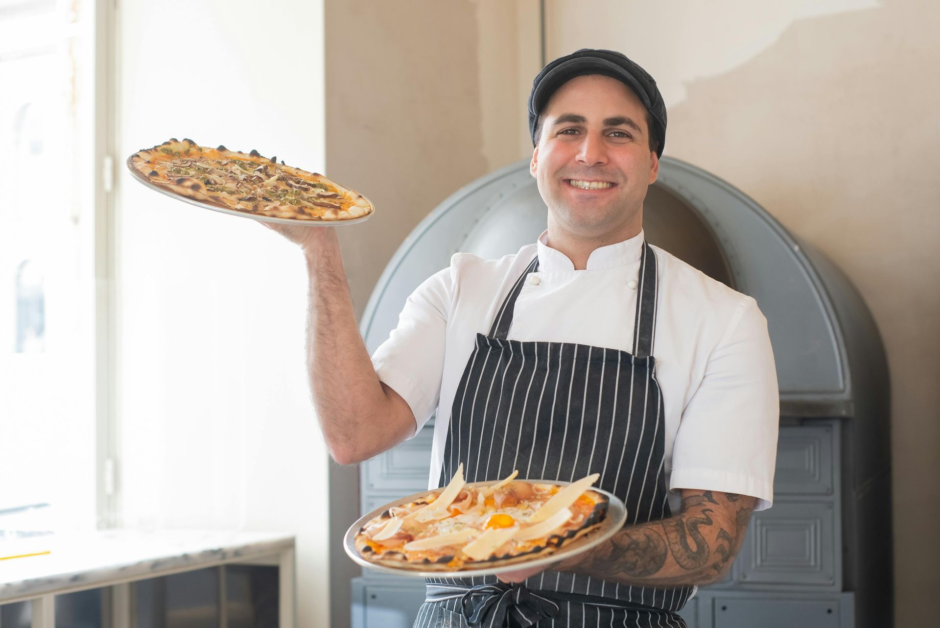 Chef holding two pizzas near a brick oven, smiling. White shirt, striped apron, black cap.