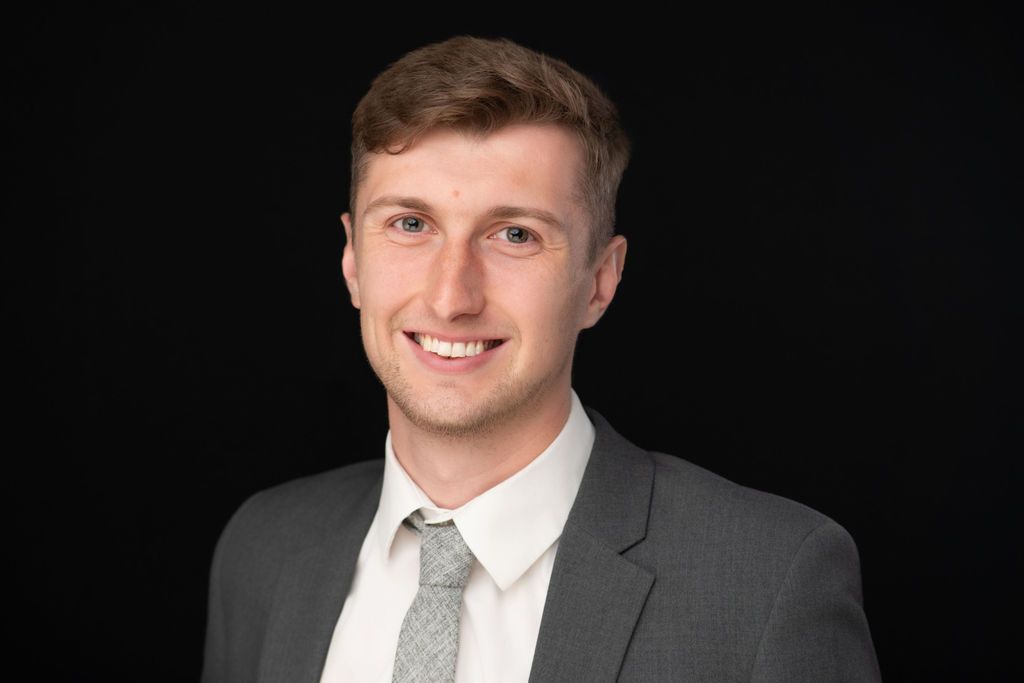 Professional headshot of a smiling person in a gray suit and white shirt against a black background