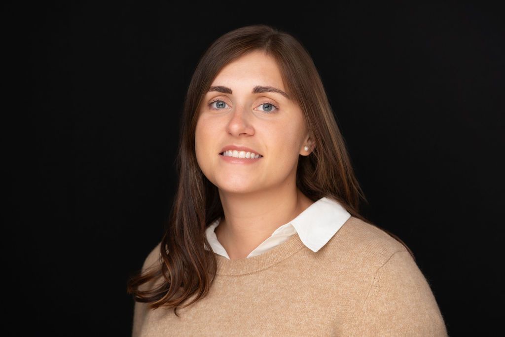 Portrait of a smiling person with brown hair wearing a beige sweater on a dark background