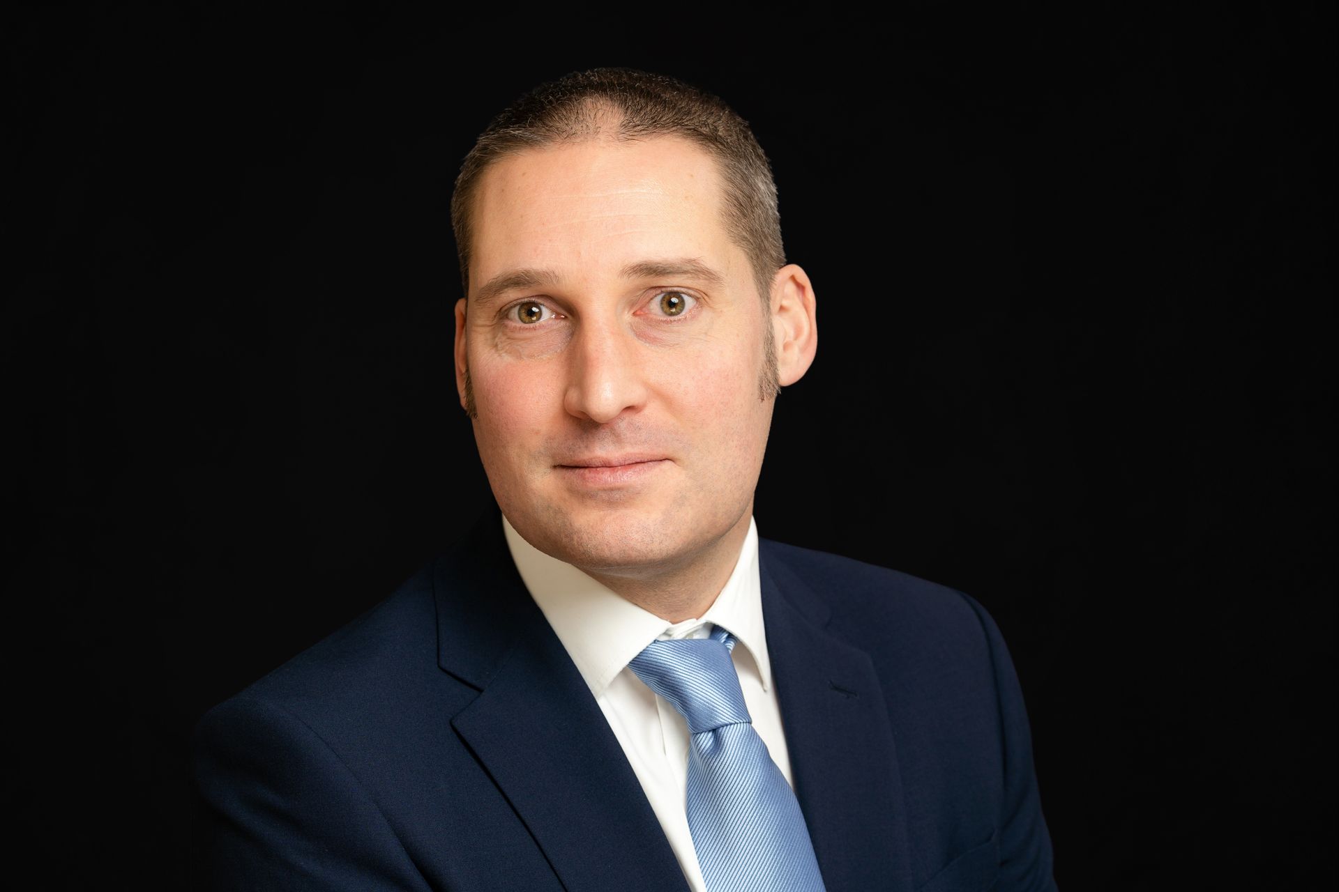 Studio headshot of a man in a dark suit and blue tie against a black background