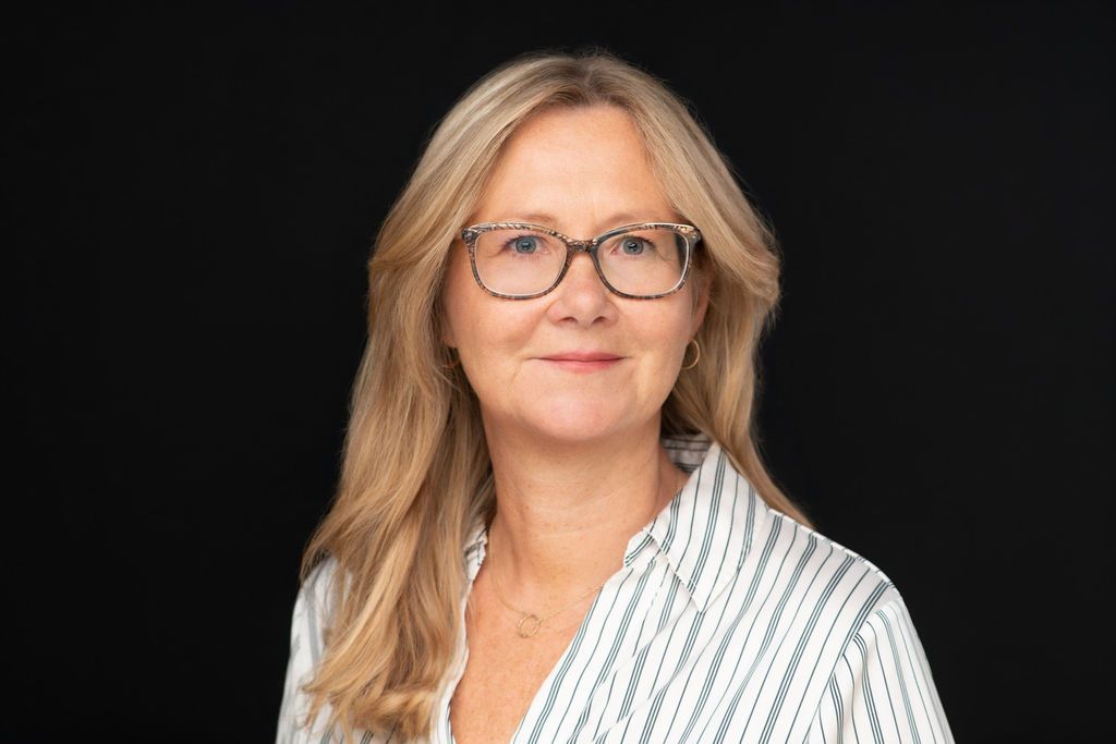 Portrait of a smiling woman with glasses and blonde hair against a dark background