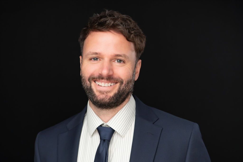 Smiling man in a navy suit, white shirt, and tie against a black background