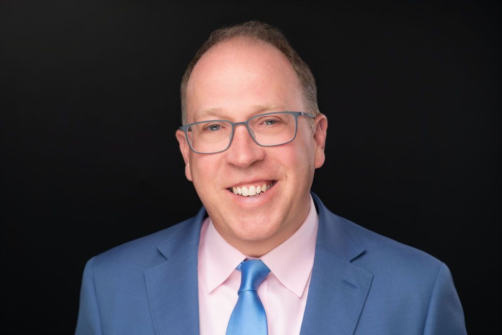 Smiling person in a blue suit and pink shirt against a black background