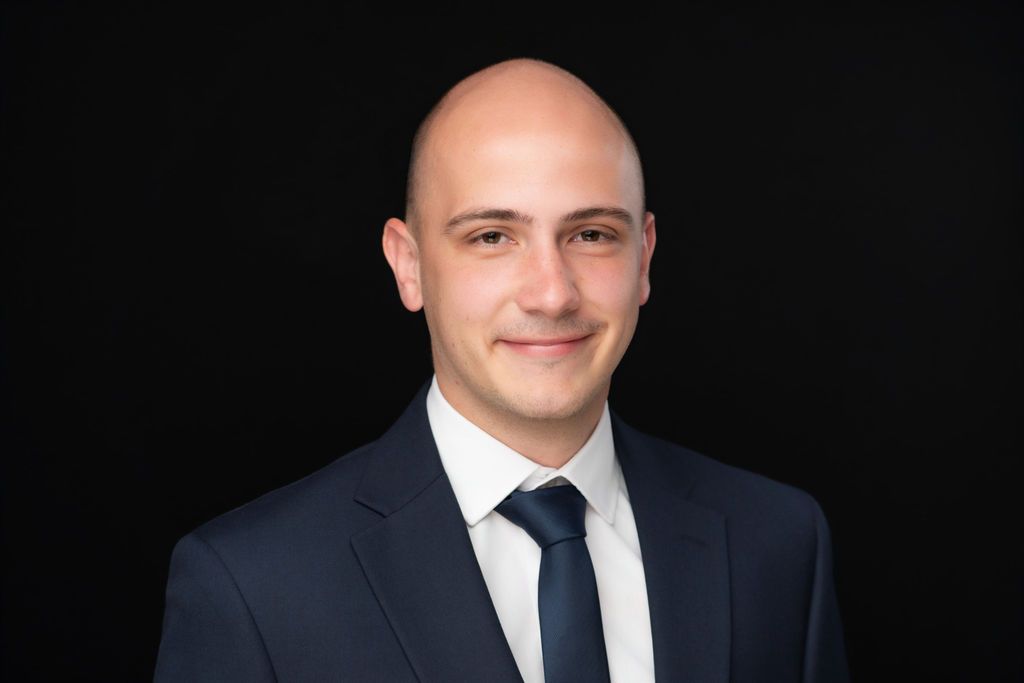 Studio portrait of a bald man in a dark suit, white shirt, and tie, smiling against a black background
