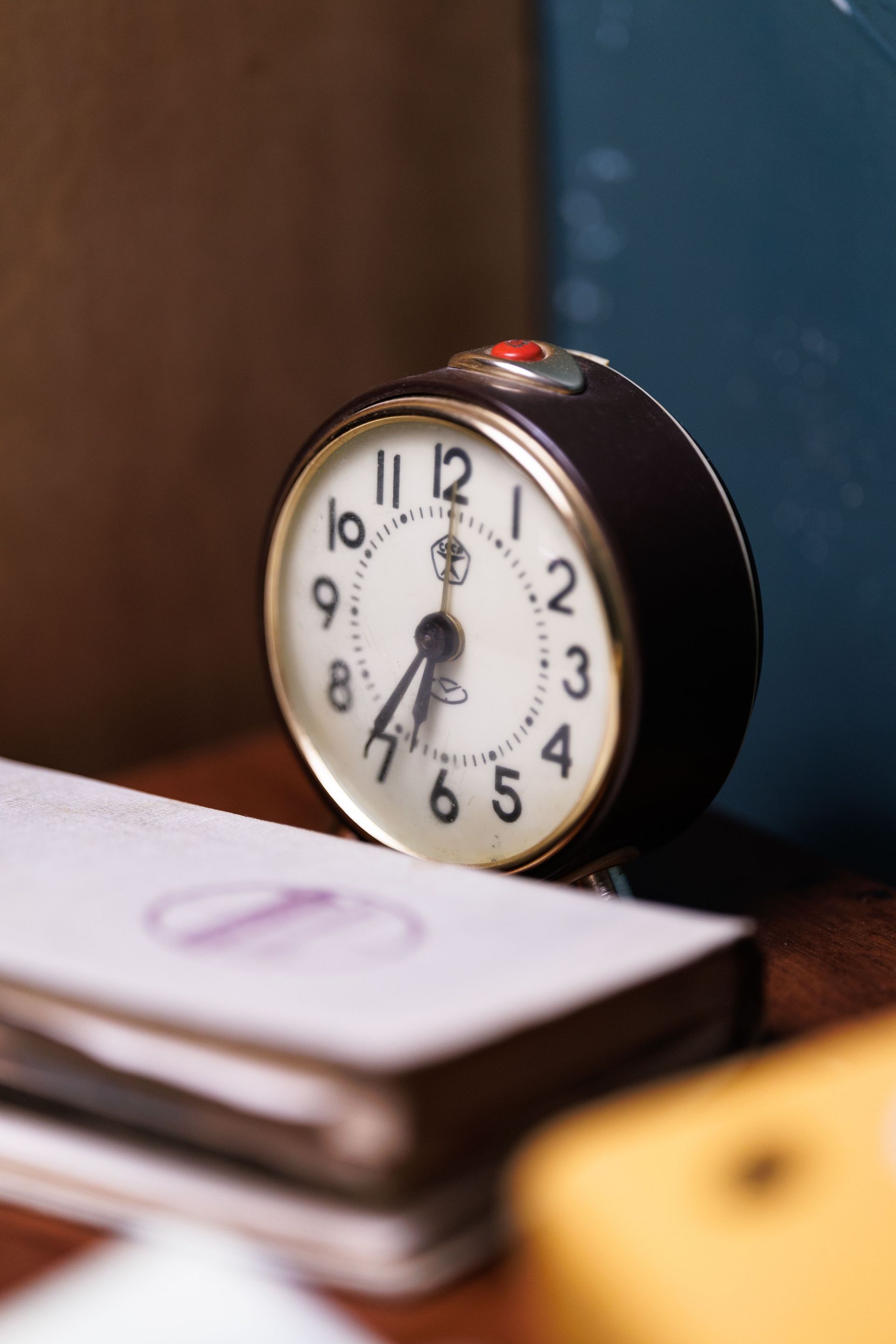 Small black alarm clock on a stack of papers beside a teal wall.