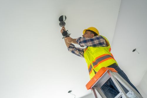Electrical contractor installing ceiling light fixture indoors, wearing hard hat and safety gear.