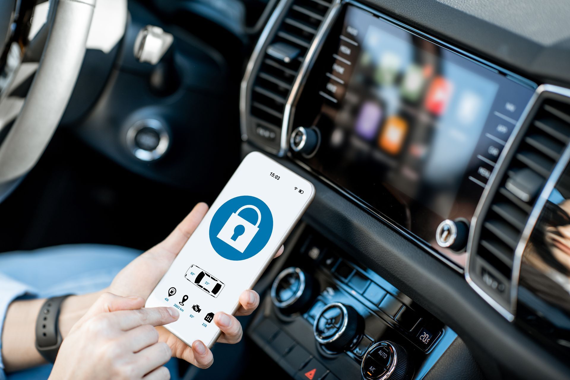 Hand holding smartphone with lock icon inside a car interior near the dashboard and infotainment screen