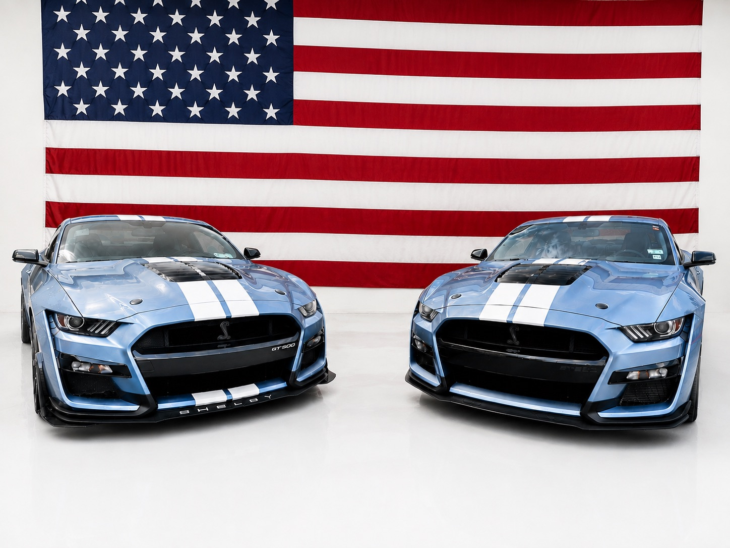 Two blue sports cars with white racing stripes in front of an American flag
