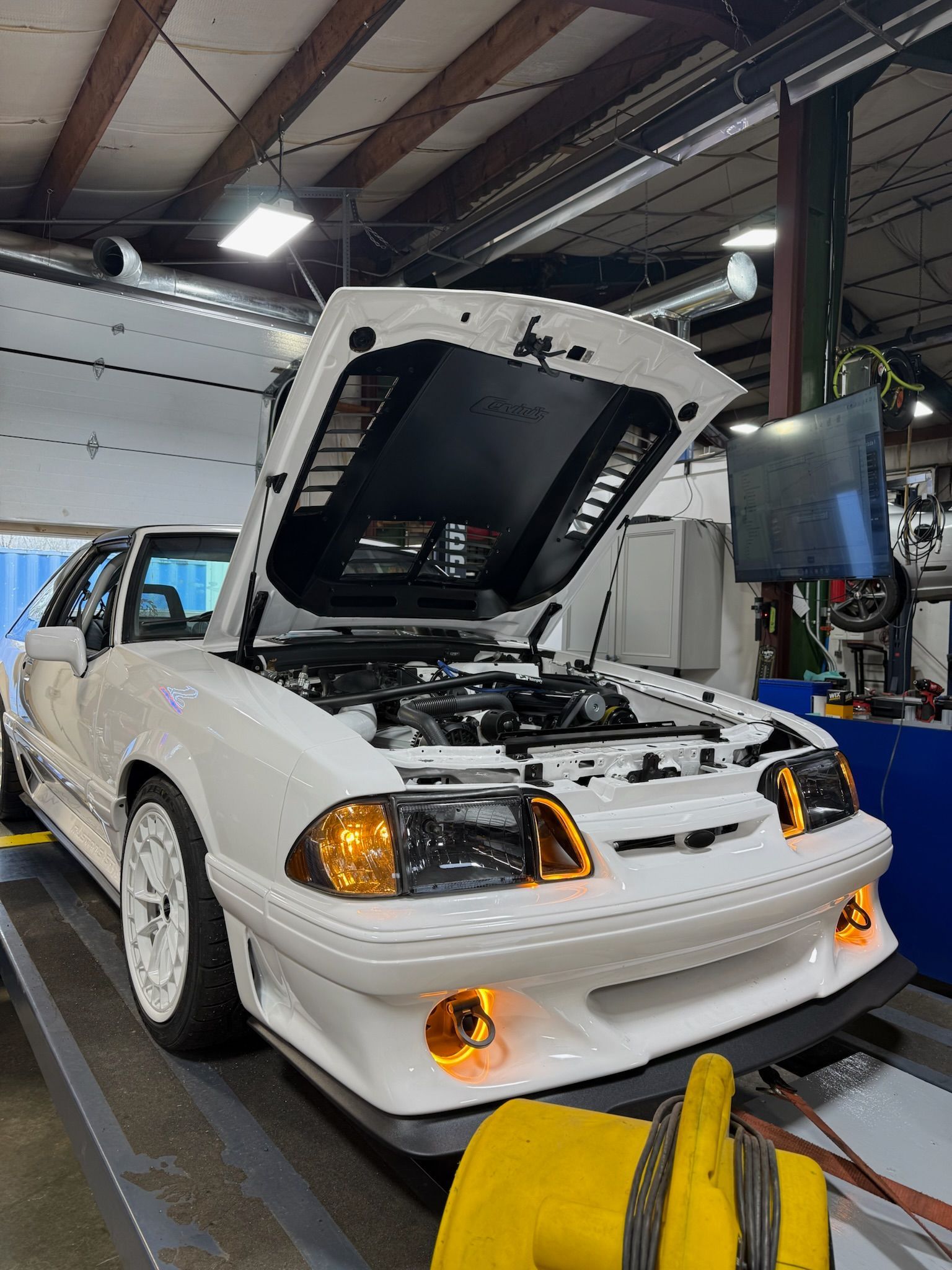 White classic car in a garage with hood open, front lights on, and engine bay exposed under bright workshop lights
