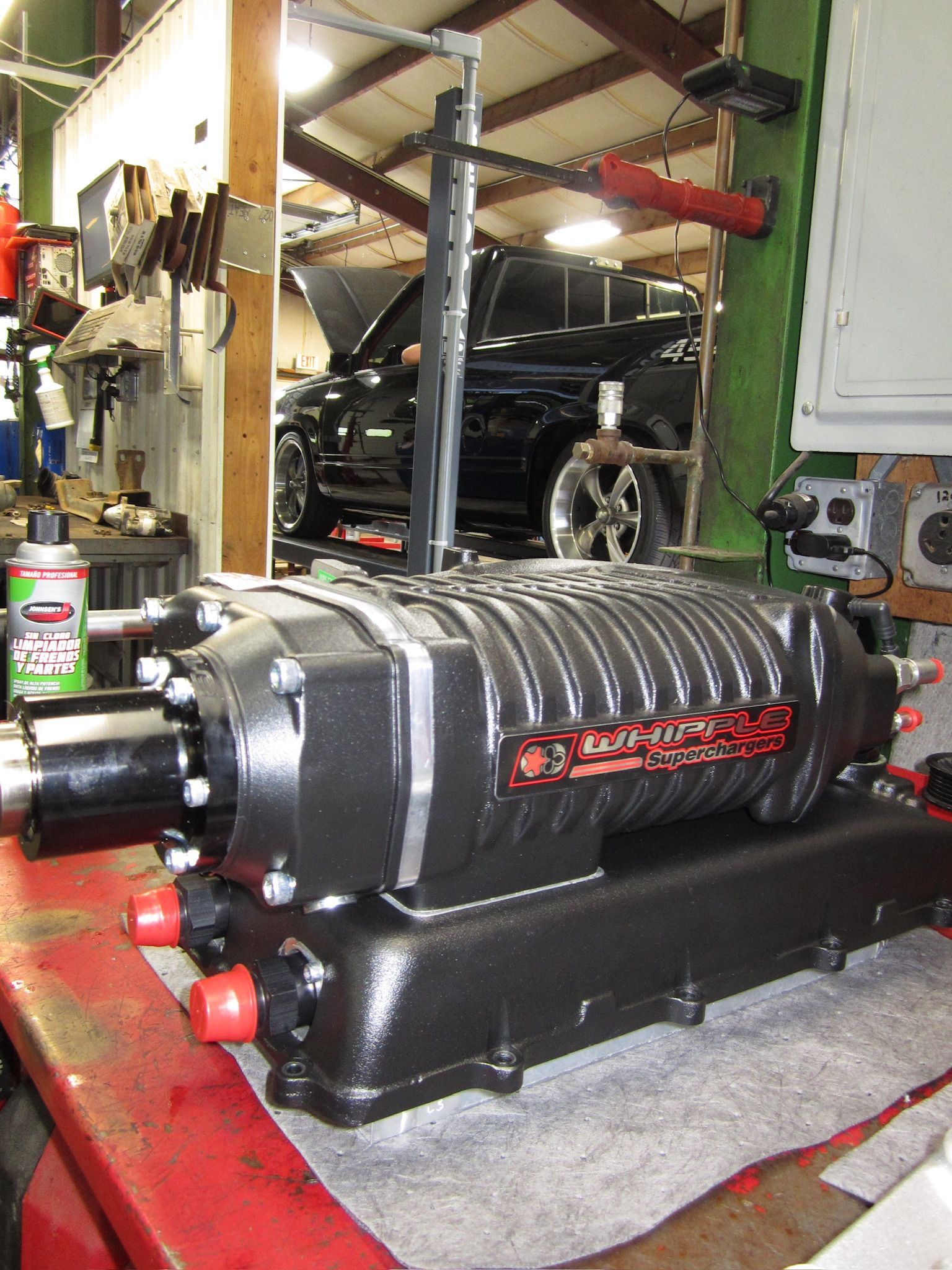 Black automotive engine with red accents on a workbench in a garage, with a car lift in the background