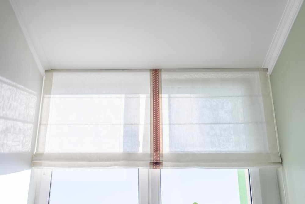 A White Roman Shade is Hanging Over a Window in a Room — Budget Screens & Awnings in South Lismore, NSW
