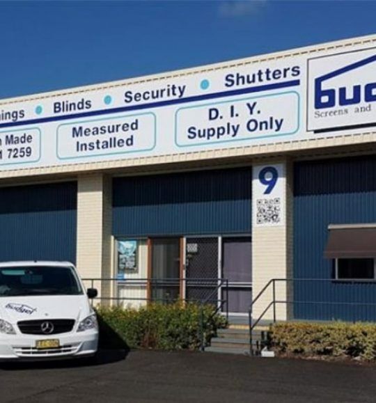 A White Car is Parked in Front of a Building That Says Shutters — Budget Screens & Awnings in South Lismore, NSW