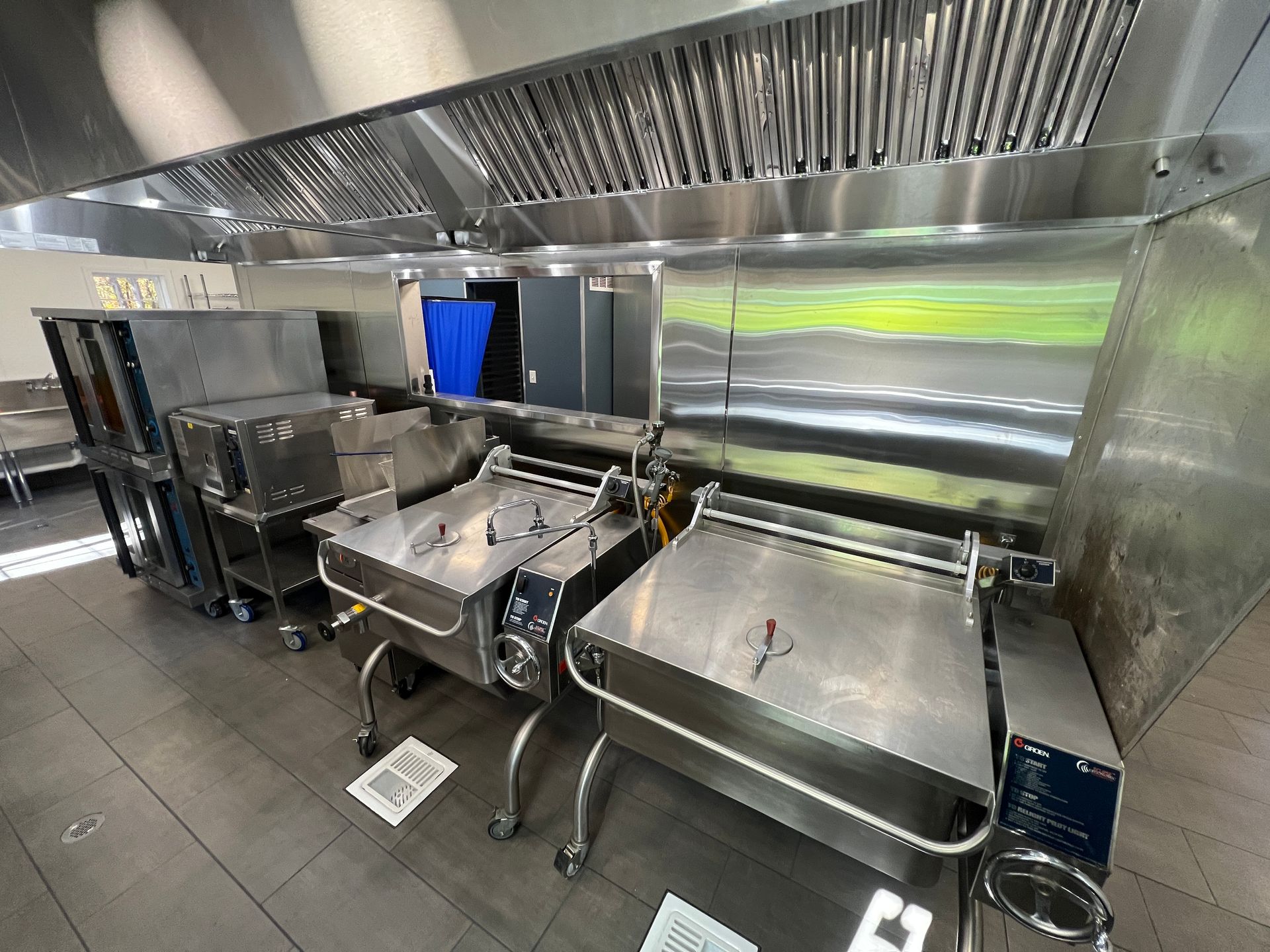 Stainless steel tilting braising pans and cooking equipment lined up under a commercial kitchen ventilation hood.