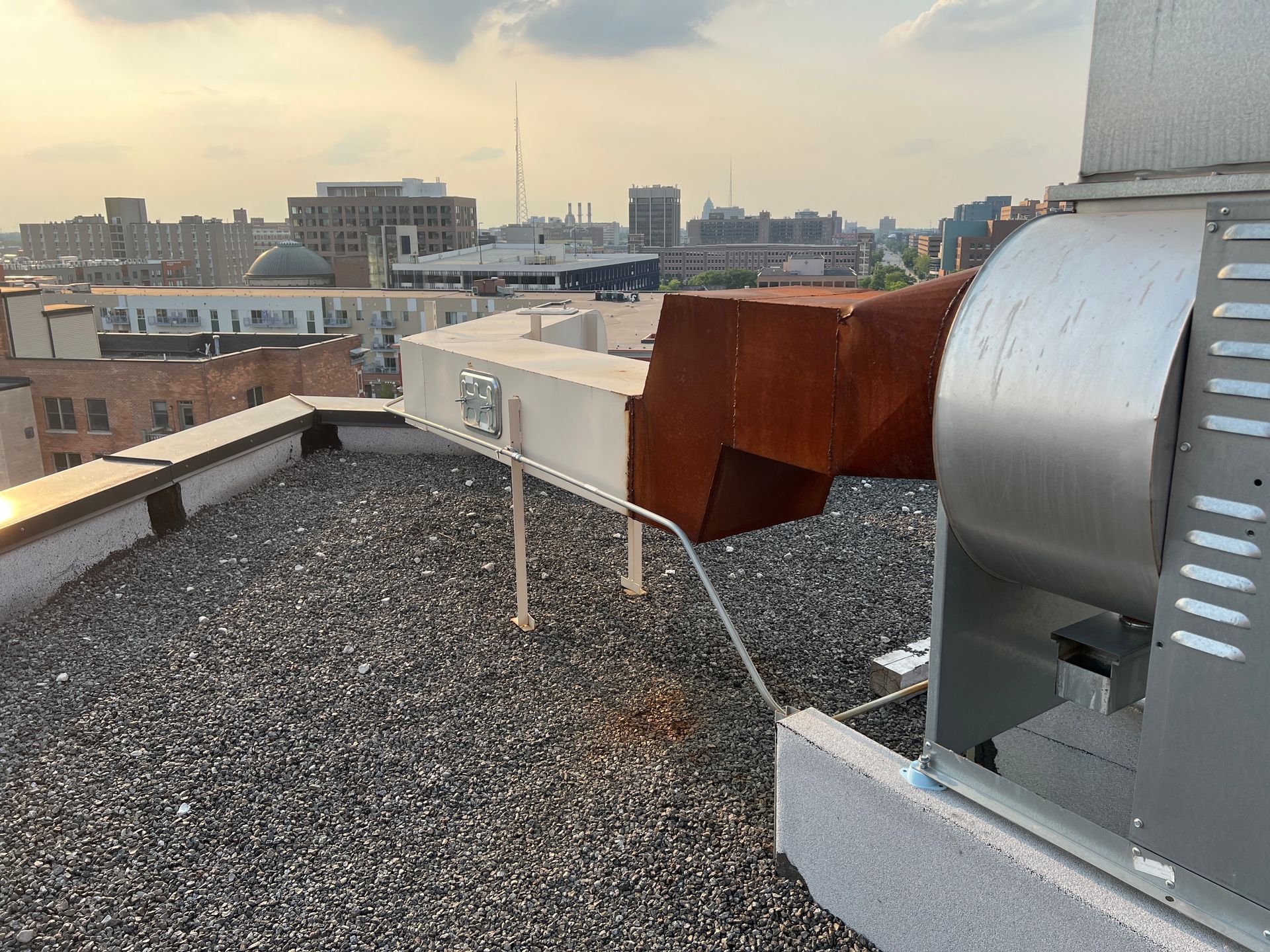 Rooftop HVAC units with white exhaust vent and rusted ductwork overlooking a city skyline.