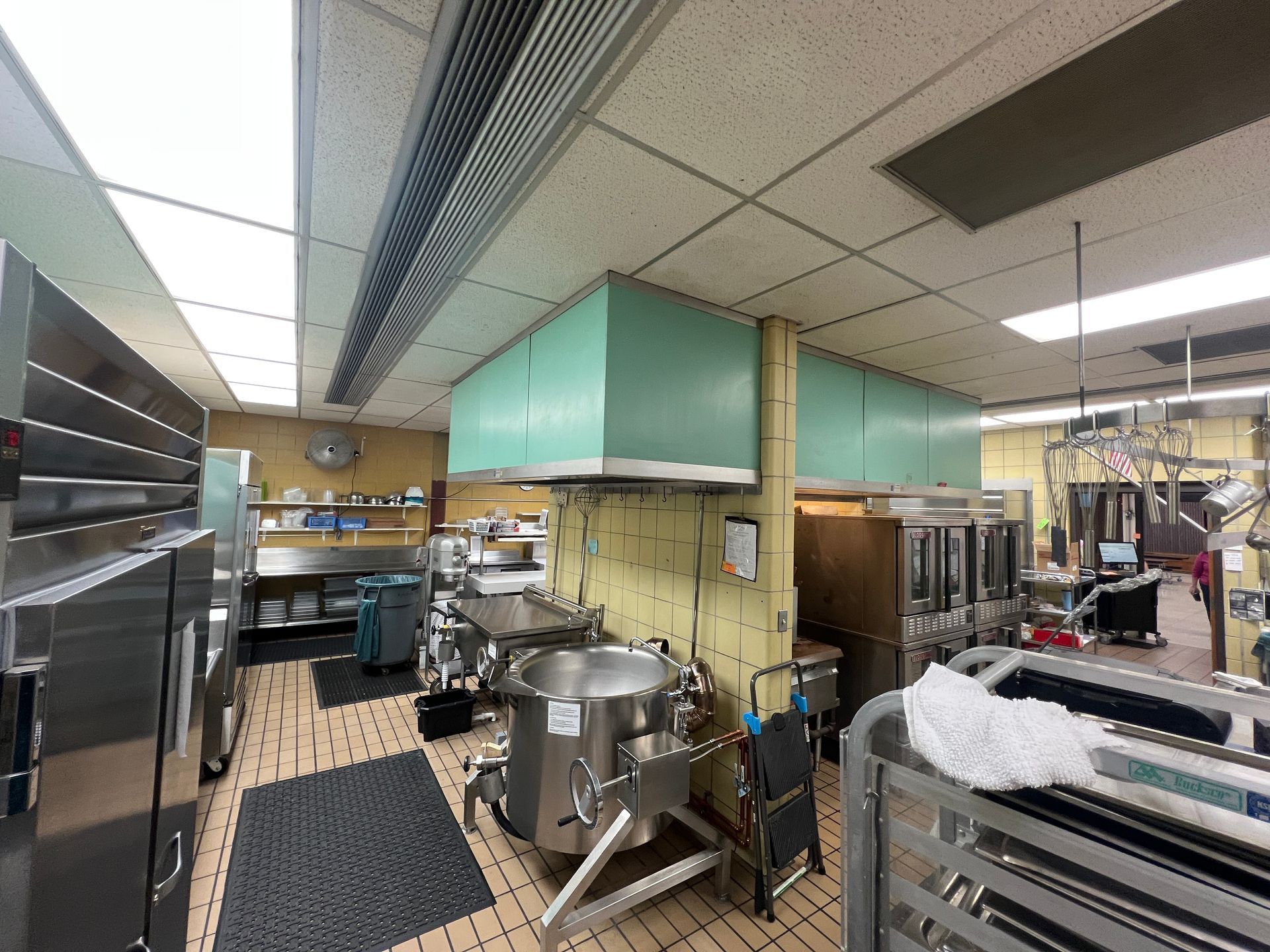 Commercial kitchen with stainless steel equipment and large pot, under green ventilation hoods.