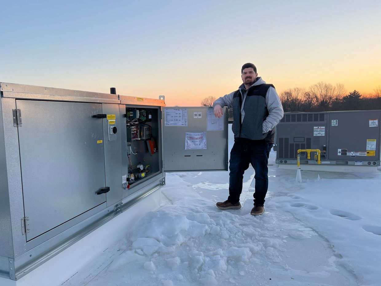 Person standing on a snowy rooftop at sunset beside an open electrical unit.