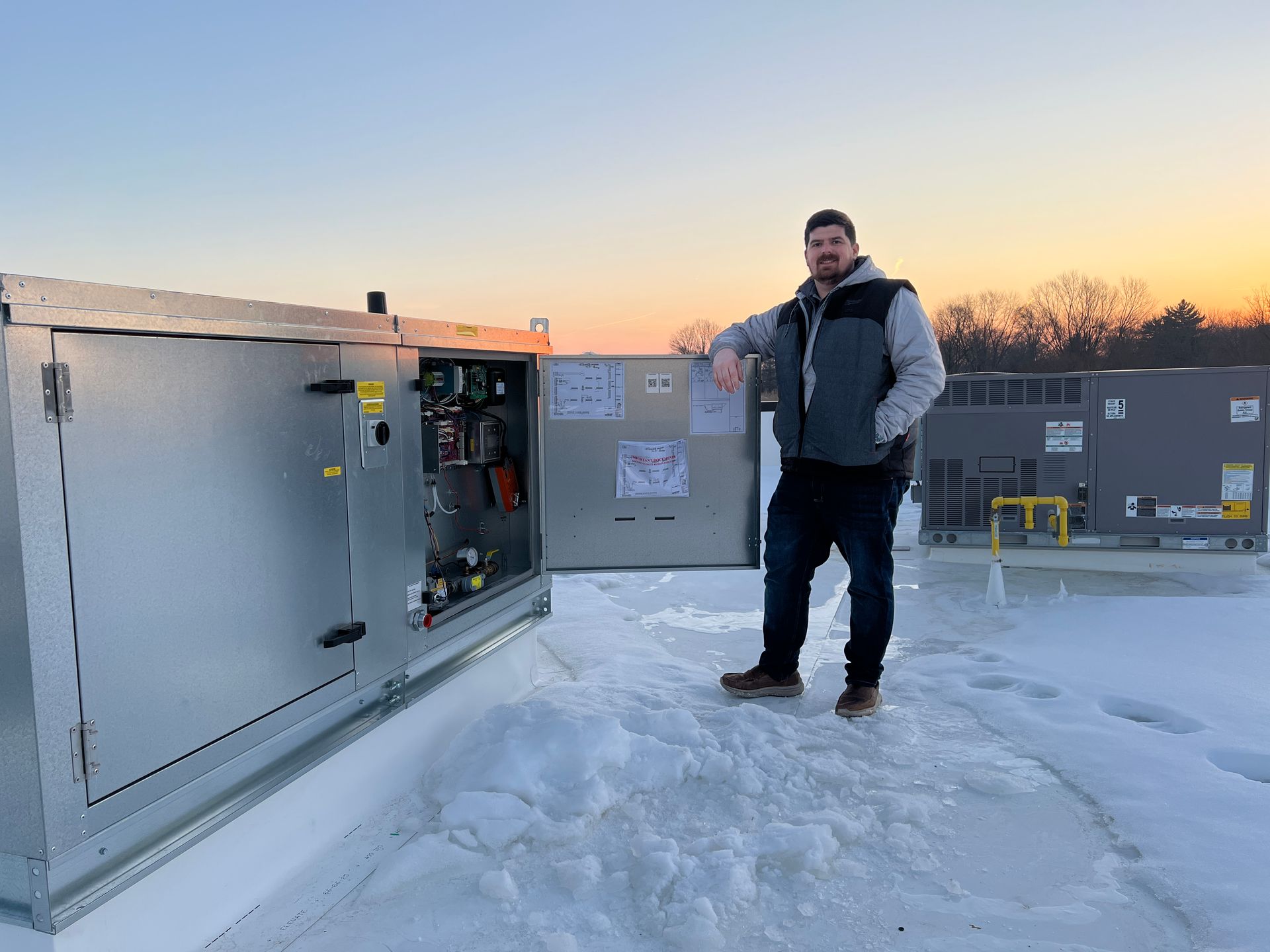 Person standing on a snowy rooftop at sunset beside an open electrical unit.