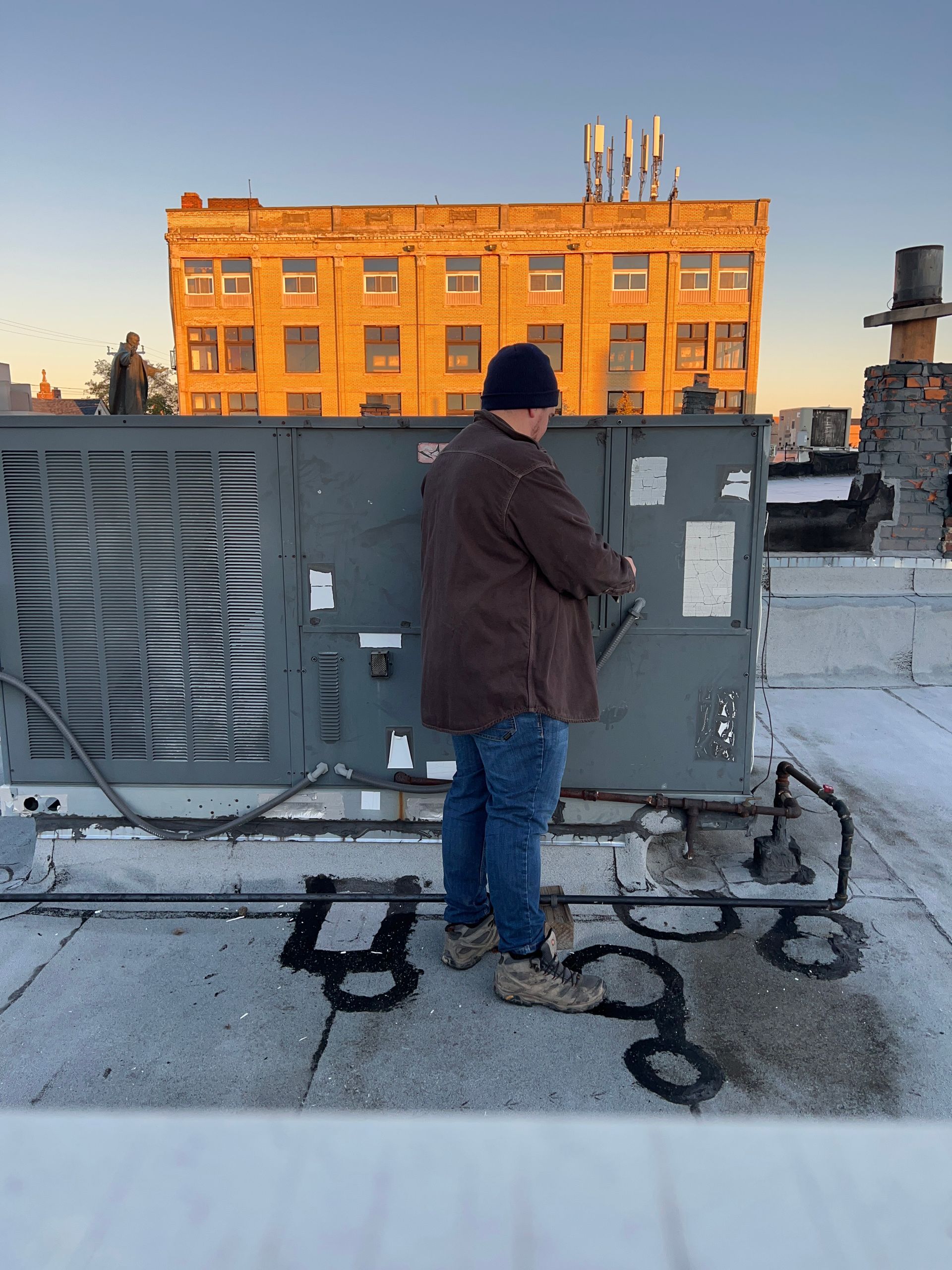 A person in a brown jacket and blue jeans services an HVAC unit on a rooftop with a yellow building in the background.