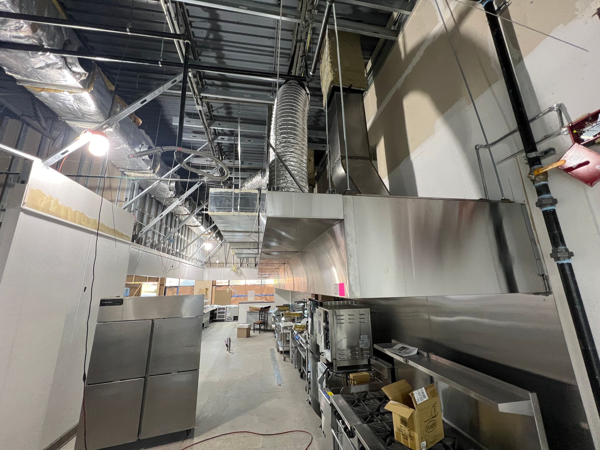 A commercial kitchen under construction with stainless steel ventilation hoods, ductwork, and equipment in a bright space.