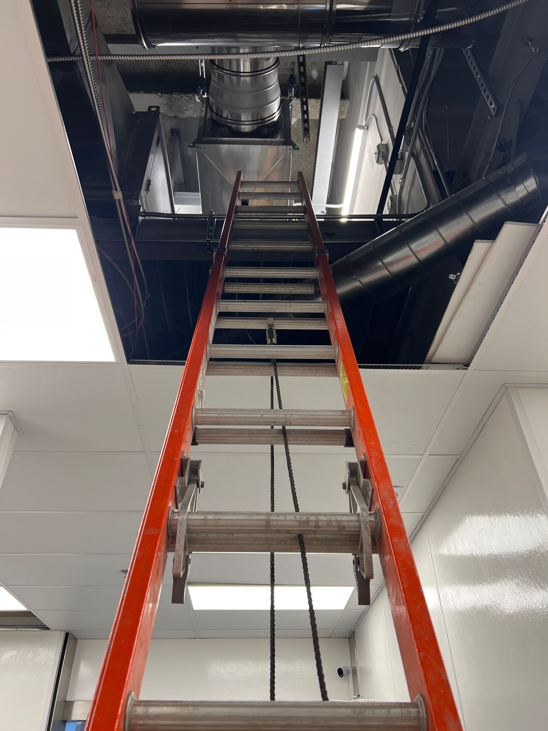 An orange stepladder positioned vertically, leading up into an open ceiling space with exposed ductwork and lighting.