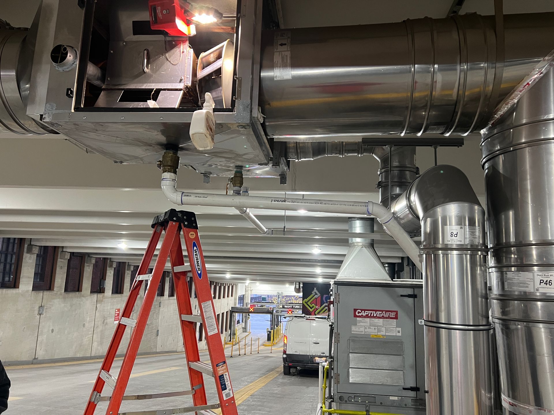 An orange step ladder stands in a parking garage under HVAC ductwork being serviced, with a work vehicle visible behind.