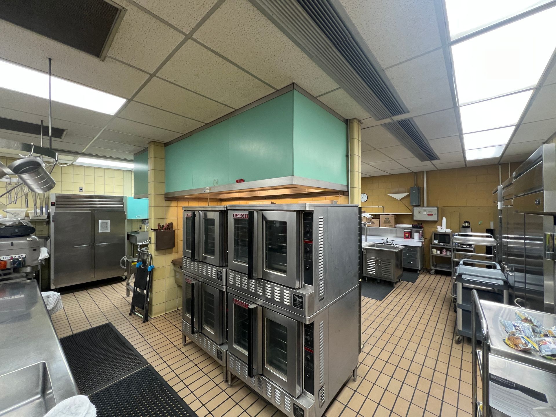 A commercial kitchen featuring a large double-stacked industrial oven under a teal exhaust hood on a tiled floor.