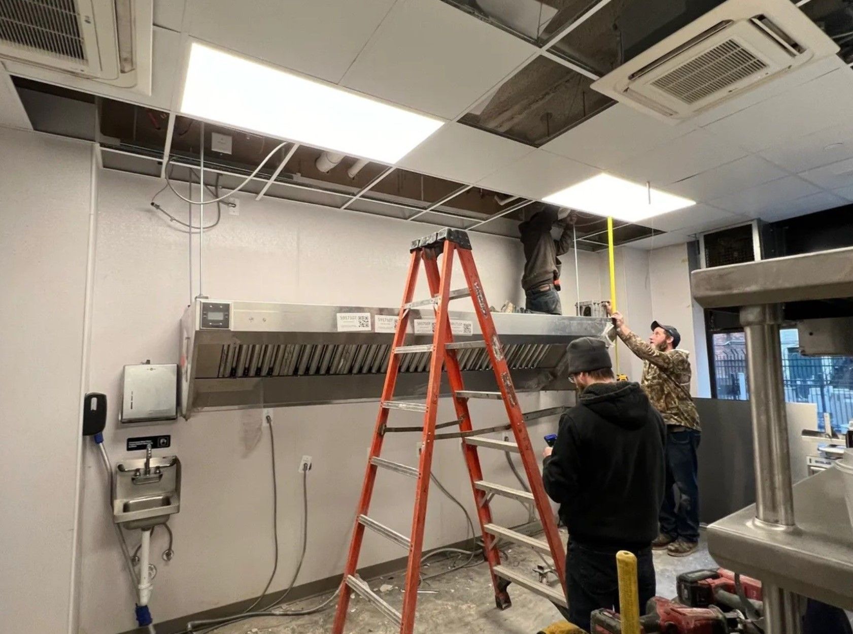 Construction workers installing a kitchen exhaust hood. One on ladder, others measuring, ceiling open, stainless steel hood installed.