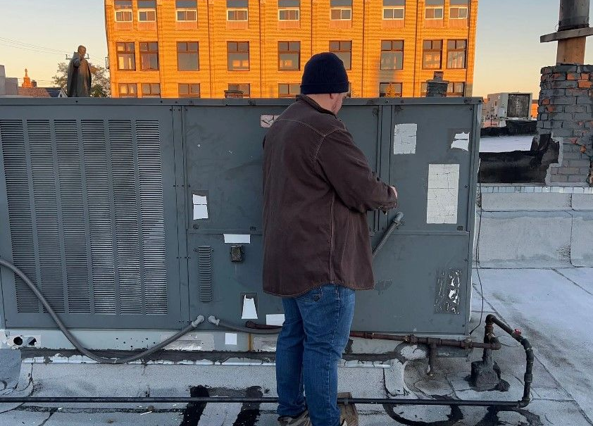 Person in brown jacket working on a rooftop HVAC unit with building in the background.