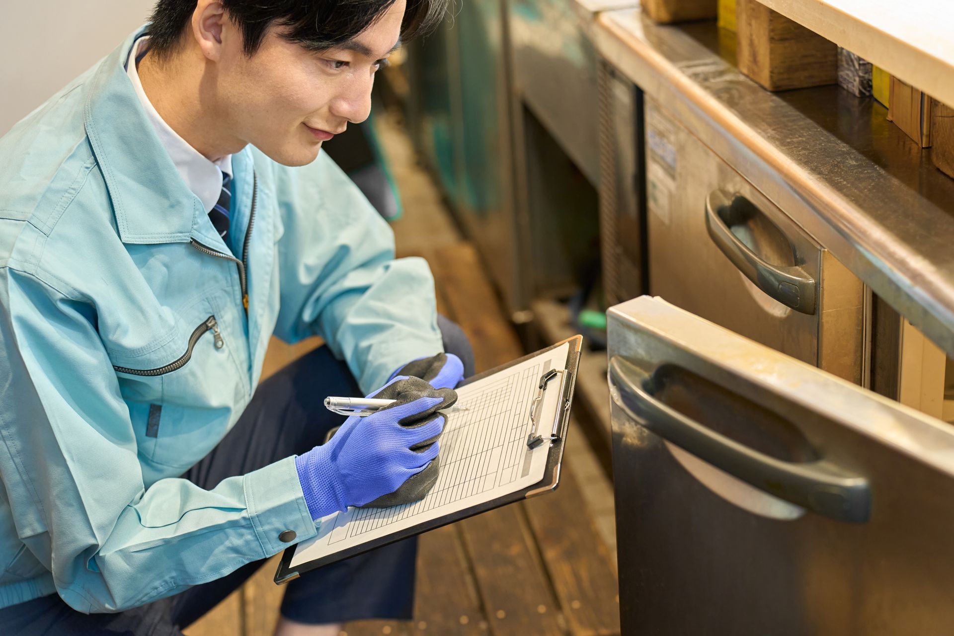 Man in blue uniform inspects equipment, taking notes on a clipboard, wearing gloves.