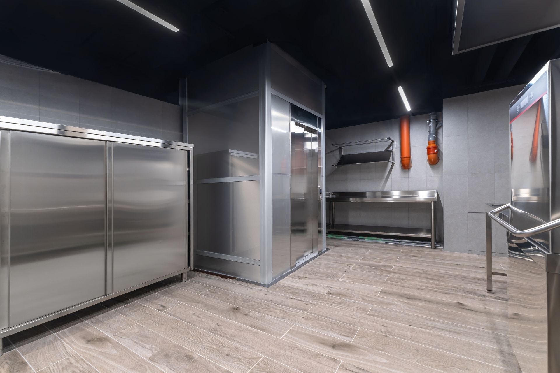 Stainless steel kitchen with elevator, refrigerator, and countertop on wooden floor. Orange fire hose on grey wall.