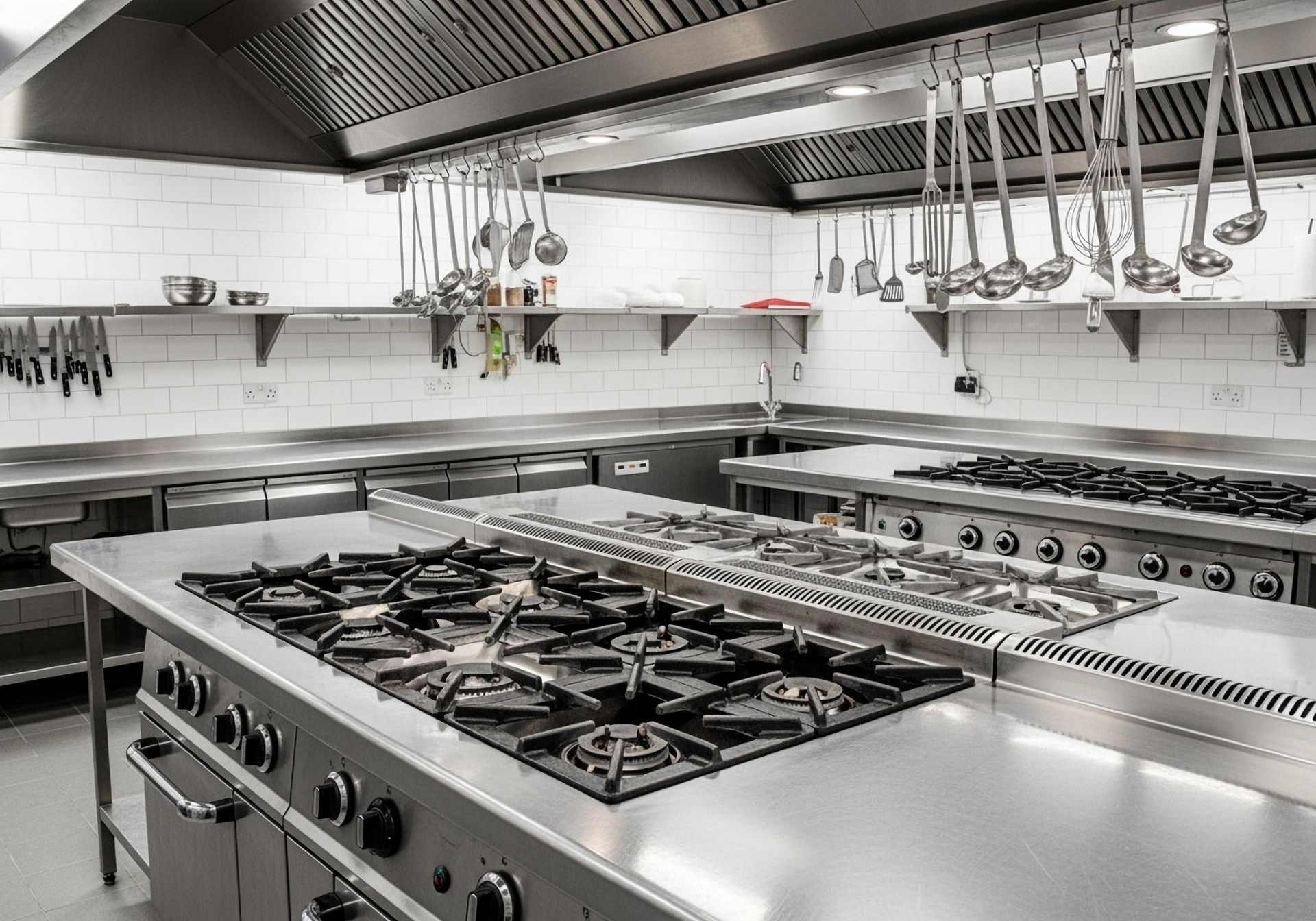 Empty stainless-steel commercial kitchen with gas stove, hanging utensils, and prep counters