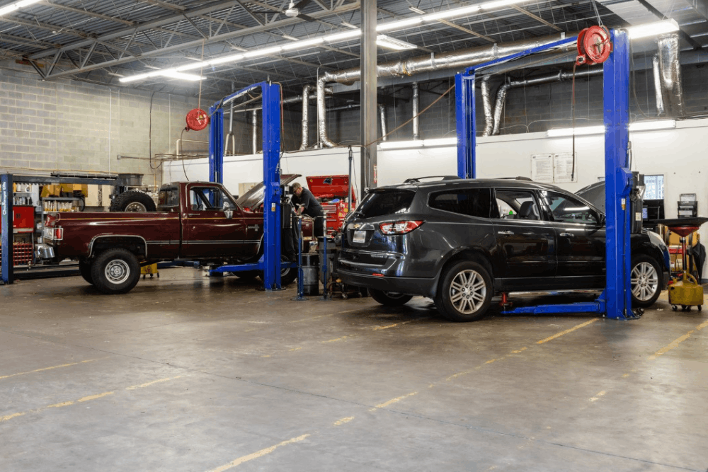 Six men in black shirts in a car repair shop; arms crossed, posing. A car is in the background.