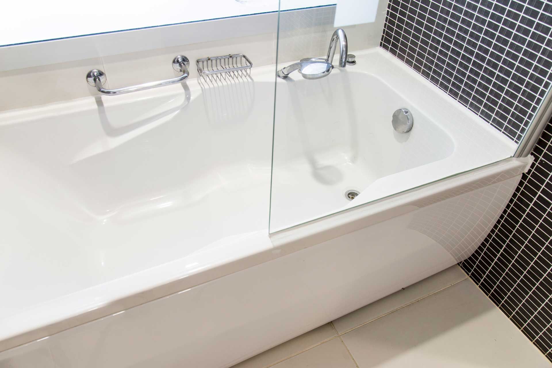White bathtub with chrome fixtures and glass shower screen in a bathroom. Black tile wall.