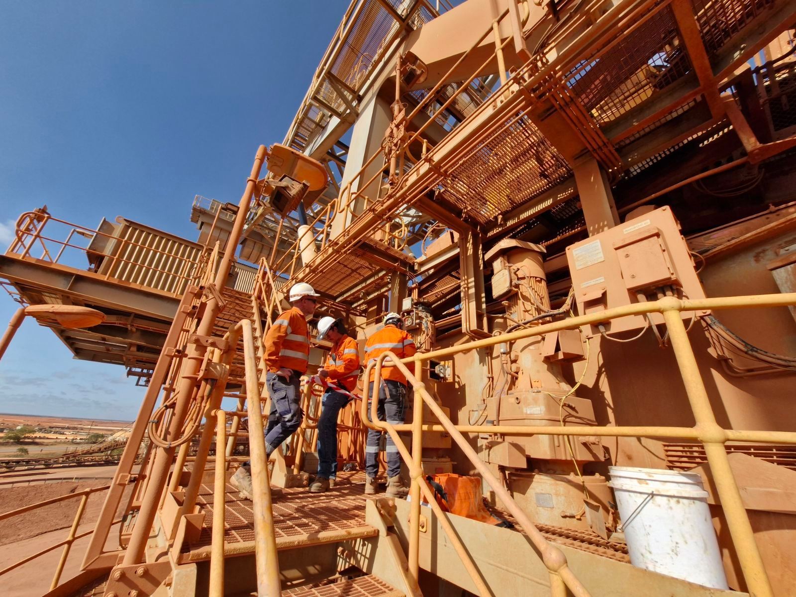 Workers in Orange Vests and Hard Hats Examining Machinery on a Brown Metal Structure Under a Blue Sky โ Projected Contracting Electrical Services In Portsmith, QLD