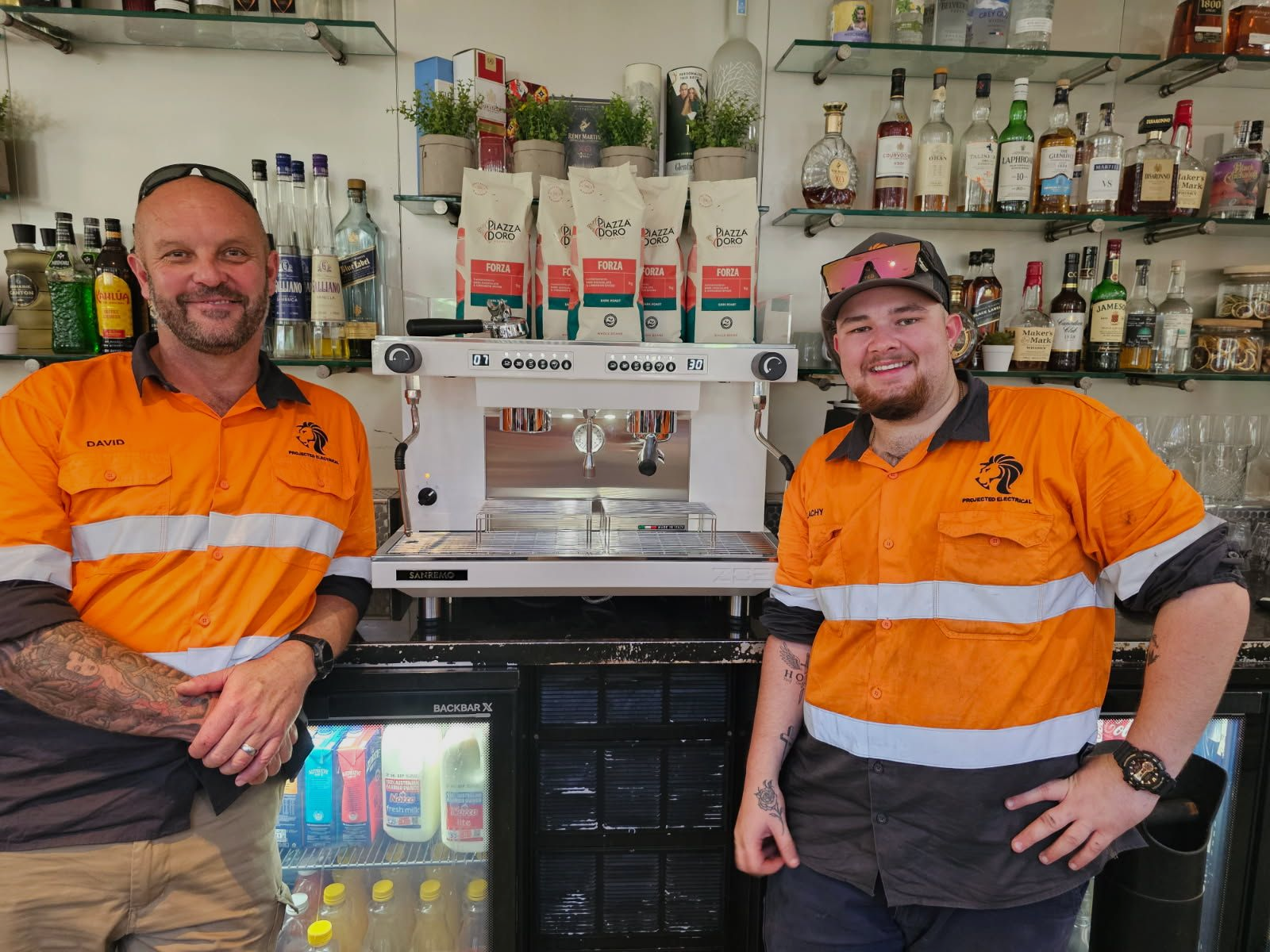 Two People in Orange Work Shirts Stand Behind a Bar — Projected Contracting Electrical Services In