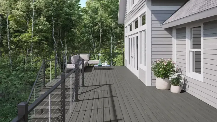 Wooden deck walkway beside a gray house, overlooking a forested hillside with potted plants and outdoor chairs.