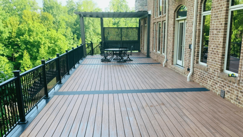 Long wooden deck with black railing beside brick house, overlooking green trees and a pergola with patio table.