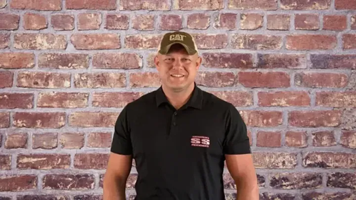 Man in a black polo and camo cap standing in front of a brick wall