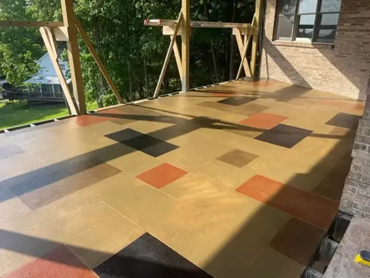 Sunlit outdoor patio with tan tiled floor and scattered brown, black, and orange squares beside a brick building.
