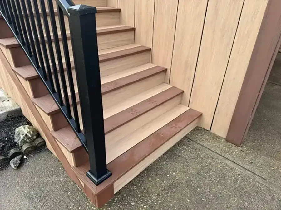 Outdoor stairs with black railing beside a beige wooden wall and concrete path