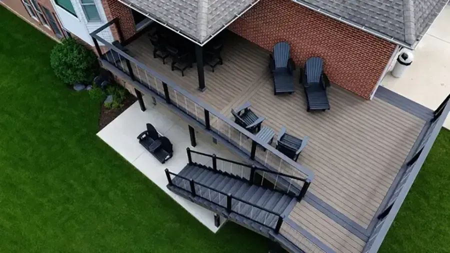 Aerial view of a backyard deck with outdoor seating, a table, and a black railing beside green lawn and brick house.