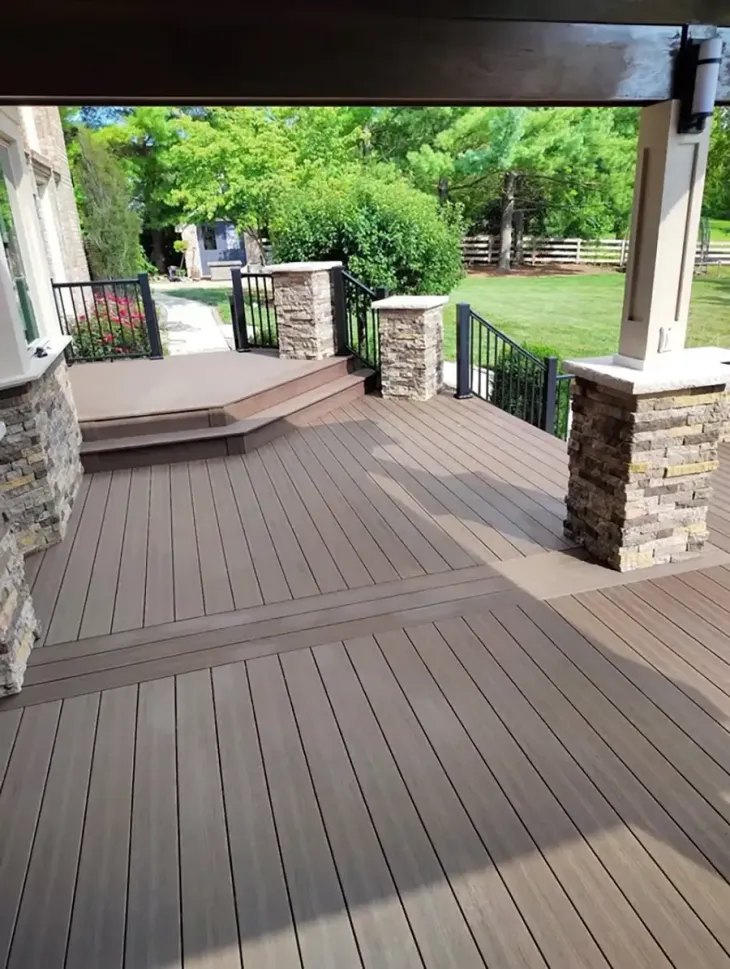 Covered wooden deck with stone pillars and railings overlooking a green backyard garden