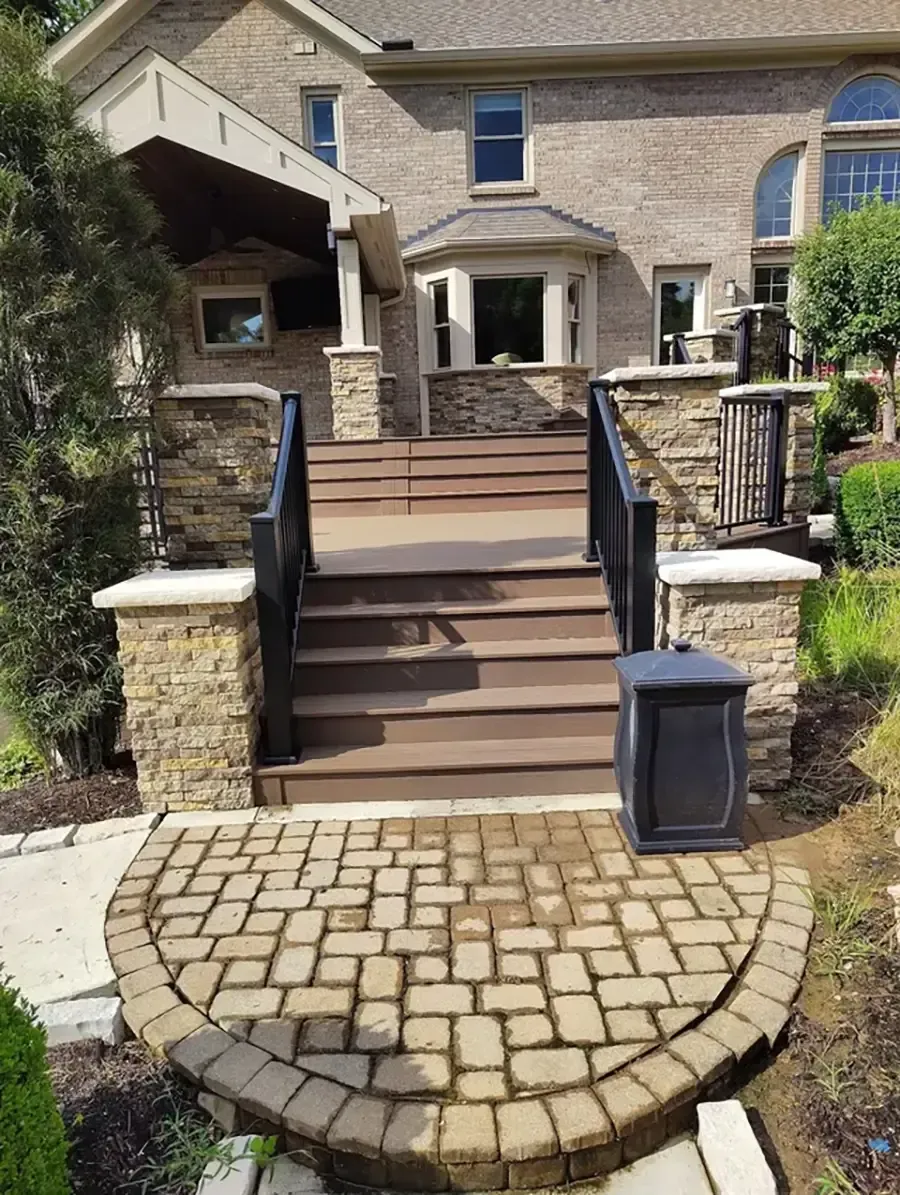 Front steps to a stone house with black railings, potted plants, and a curved brick walkway.