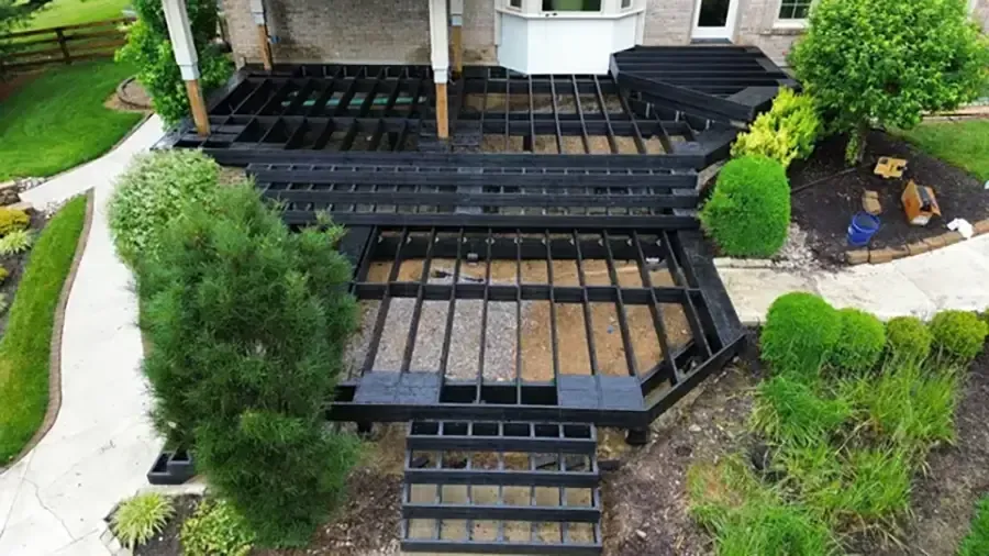 Backyard deck framing under construction beside a house, with black joists and surrounding greenery